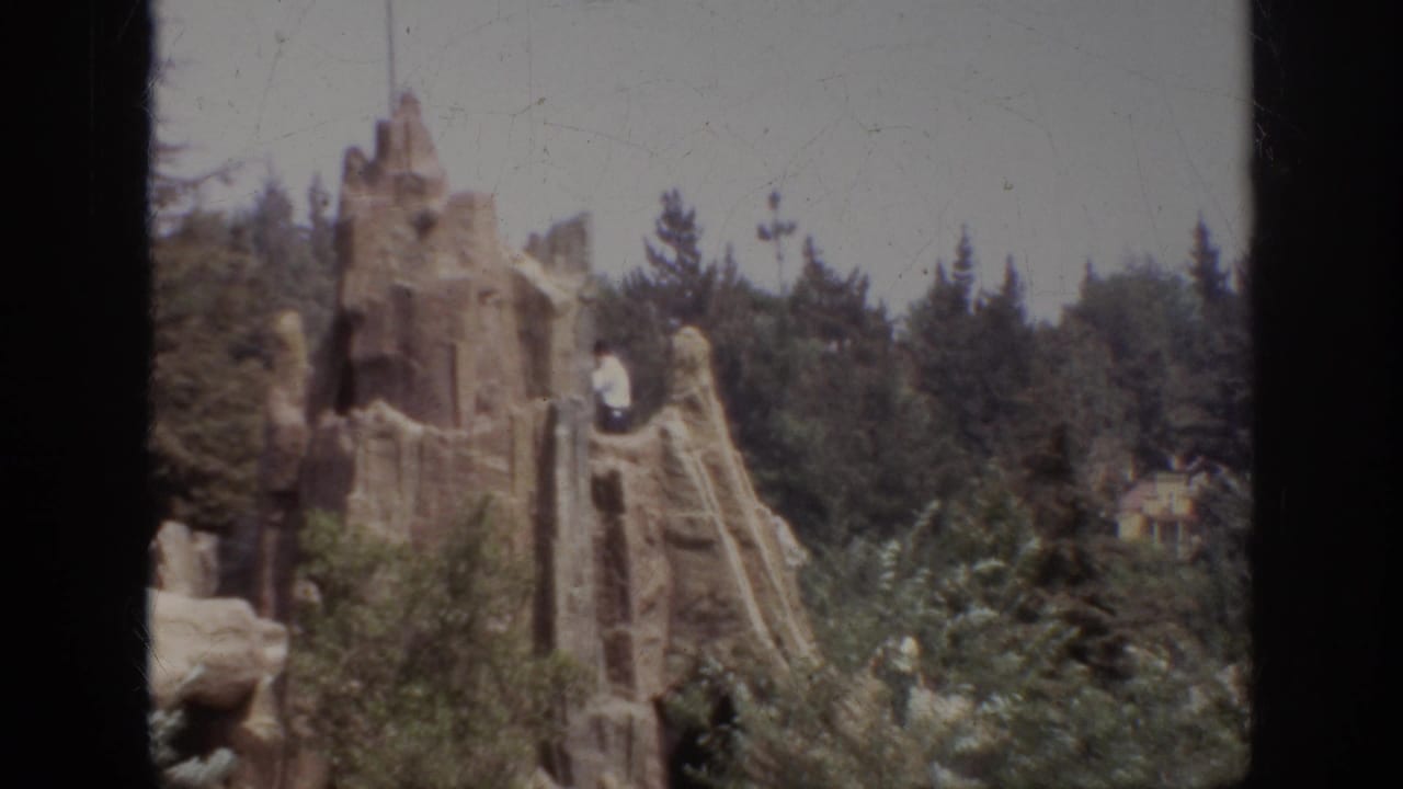 1962 Anaheim home-movie frame showing workers near an artificial stone structure in a wooded attraction setting.