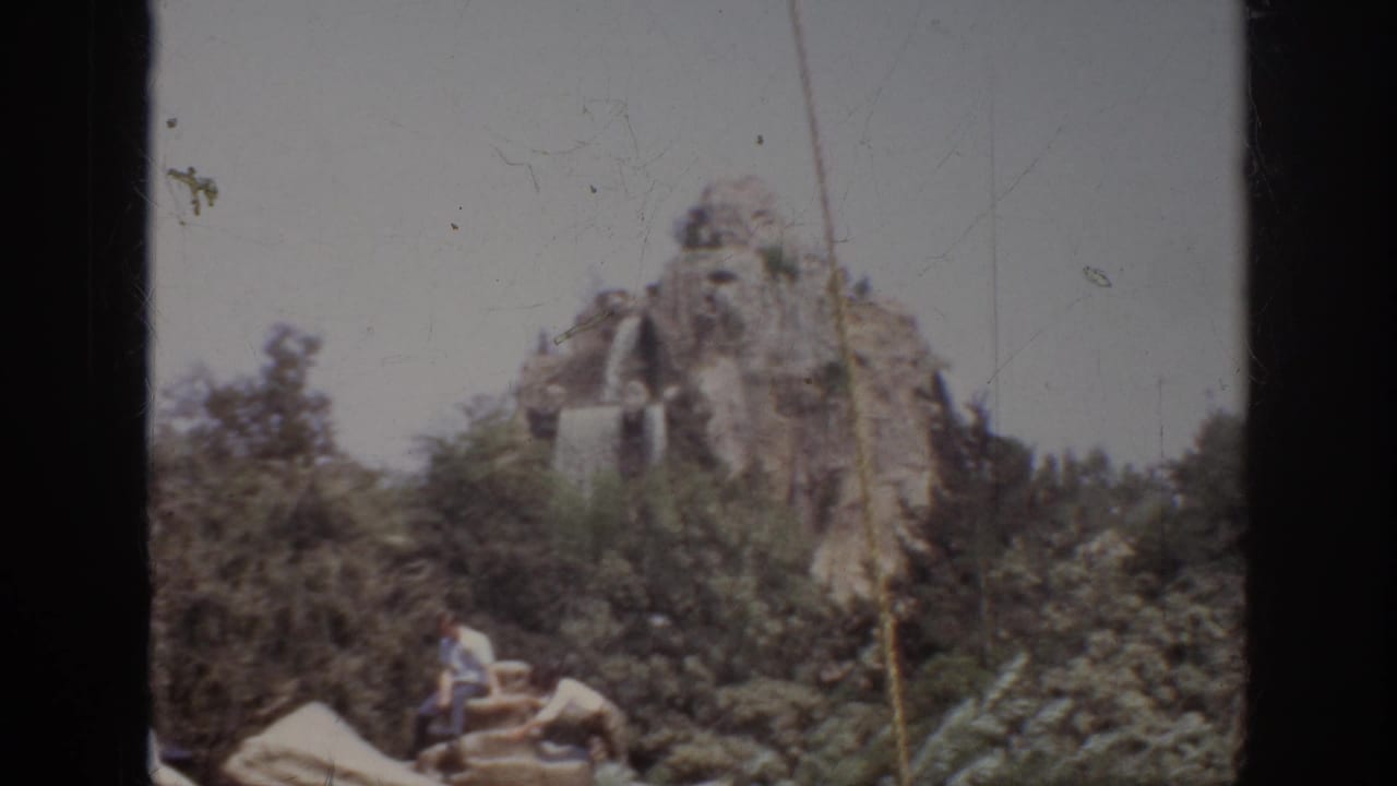 1962 Anaheim home-movie frame showing an artificial mountain, cascading water, greenery, and visitors seated near the attraction scenery.