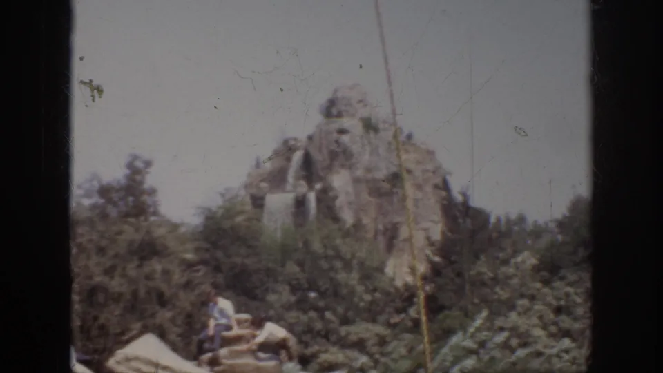 1962 Anaheim home-movie frame showing an artificial mountain, cascading water, greenery, and visitors seated near the attraction scenery.