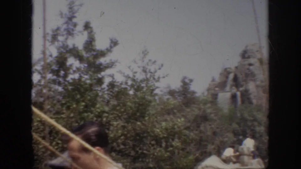 1962 Anaheim home-movie view with a foreground visitor, diagonal ropes, greenery, and distant artificial rockwork with water.