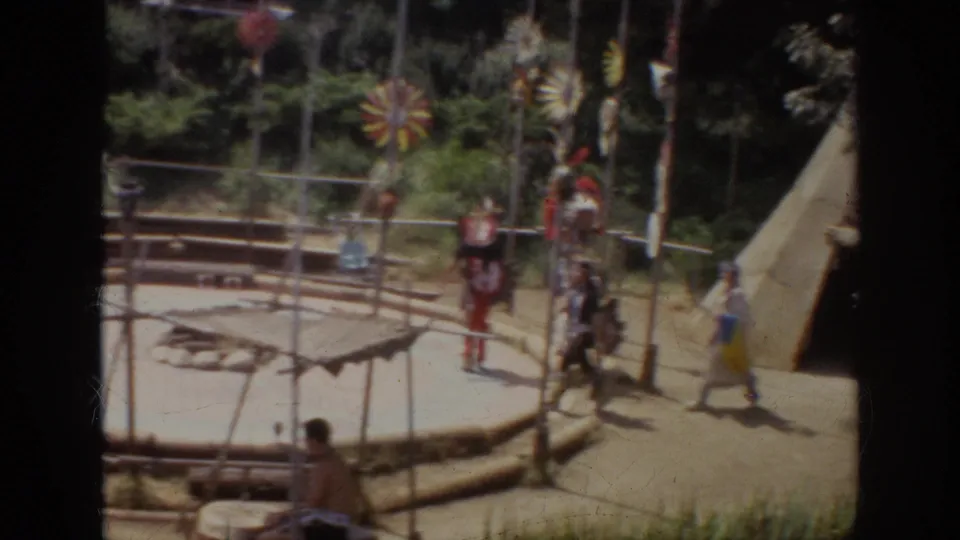 1962 Anaheim frame showing performers in Native American-inspired regalia near teepee-like structures, colorful poles, fencing, and visitors.