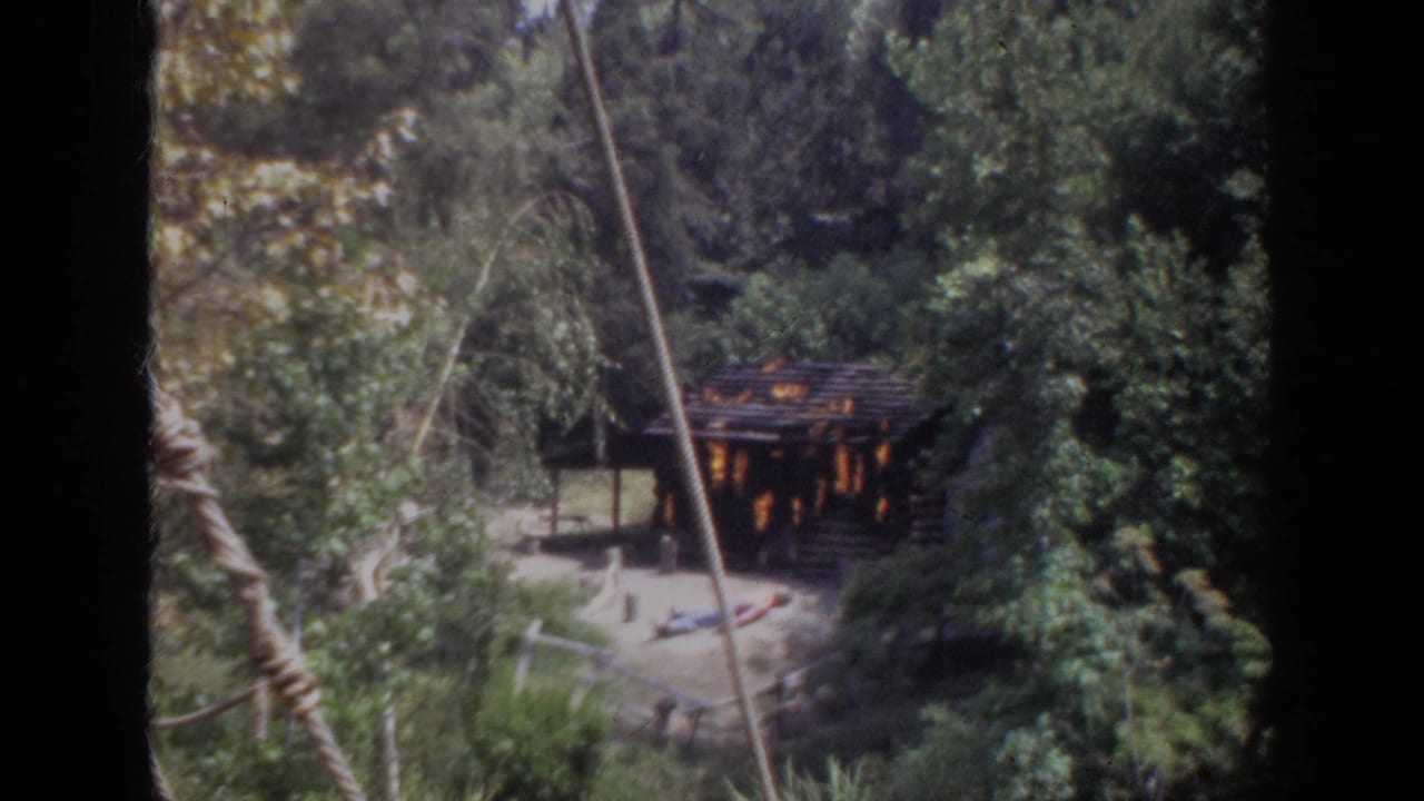1962 Anaheim 8mm frame showing a rustic cabin burning in a wooded setting, viewed from above with ropes or cables in the foreground.