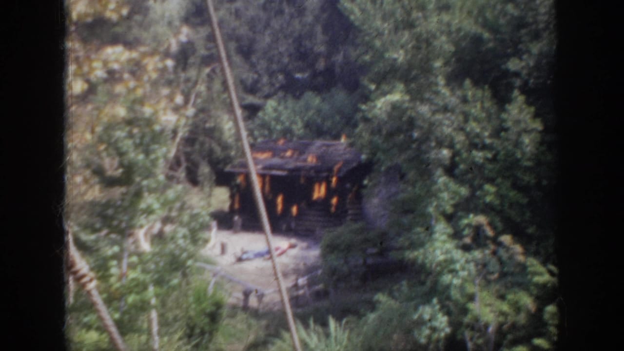 1962 Anaheim home-movie frame of a cabin fire in dense greenery, with flames visible through openings and a foreground cable crossing the view.