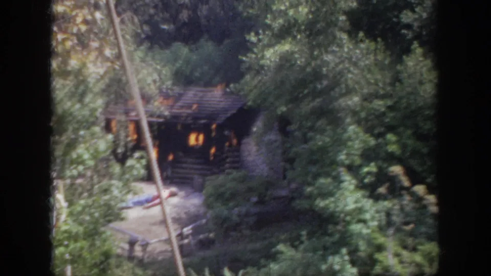 1962 Anaheim 8mm frame showing a burning rustic cabin in woods, with small human figures visible near the pale ground area below.