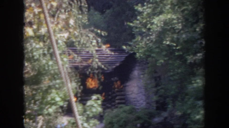 1962 Anaheim home-movie frame of flames inside a rustic cabin, partly hidden by trees and a diagonal foreground rope or cable.