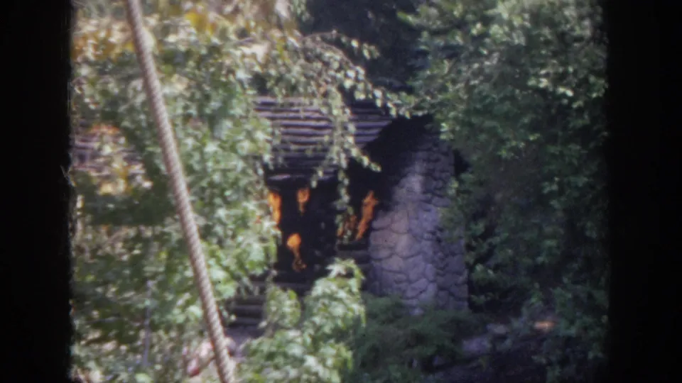 1962 Anaheim 8mm frame showing a rustic stone and log oven or wall behind dense green foliage.