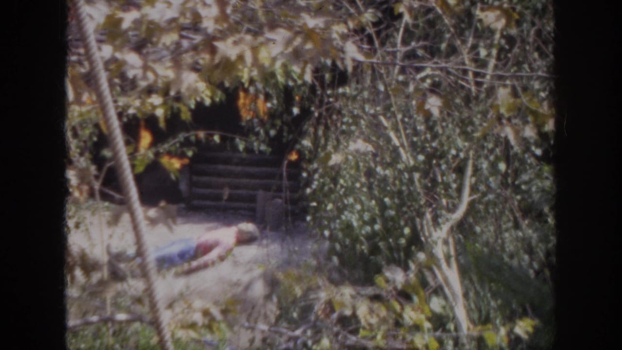 1962 Anaheim home-movie frame showing a small shack on fire with a person lying on the ground in a wooded attraction scene.