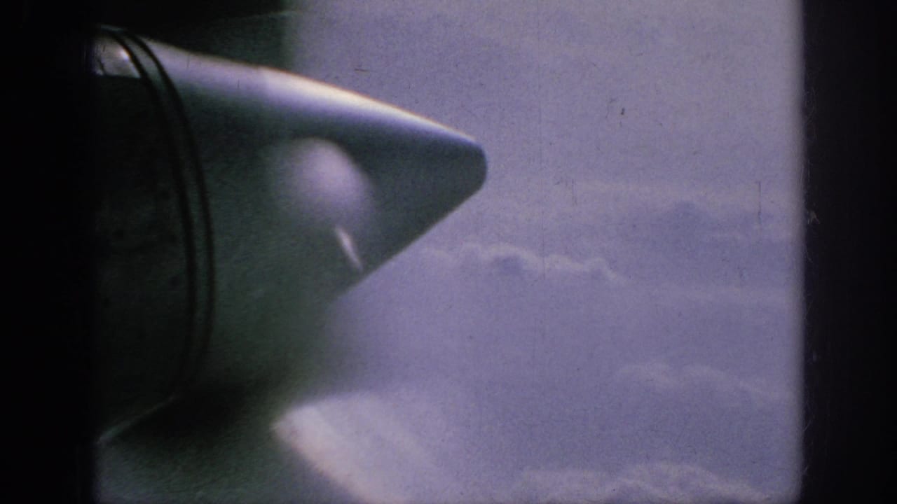 1962 8mm home-movie frame showing an aircraft wingtip through a passenger window above soft cloud layers.