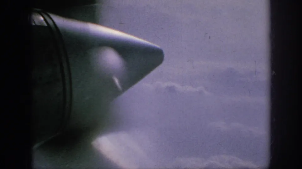1962 8mm home-movie frame showing an aircraft wingtip through a passenger window above soft cloud layers.