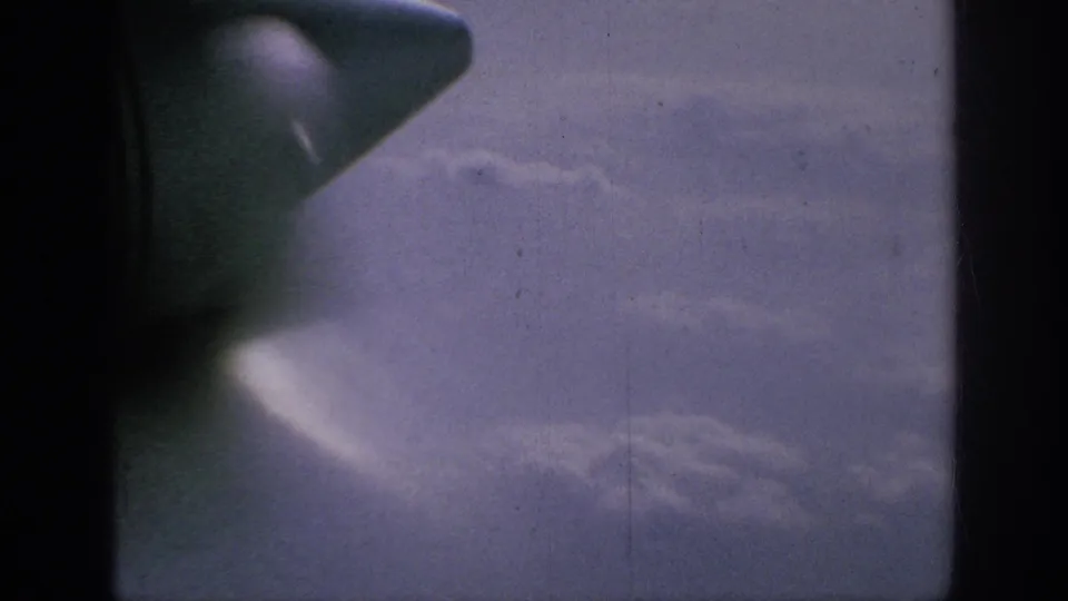Passenger-window view from 1962 8mm footage, with the wingtip near a dark divider and clouds beyond.