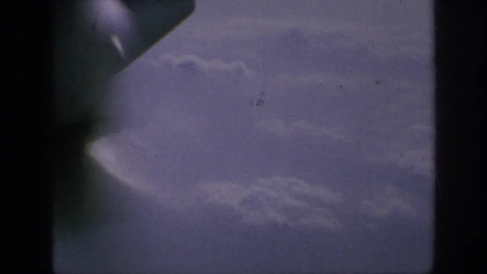 8mm frame from an airplane window, showing a partial wingtip and cloud layers in a blue-purple sky.