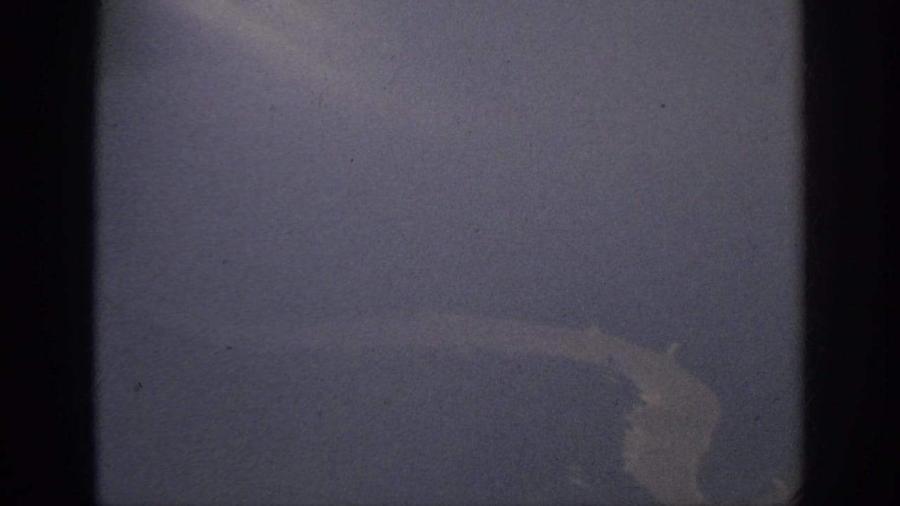 1962 Anaheim-area 8mm frame showing a hazy aircraft-window view of pale water or shoreline against darker terrain.