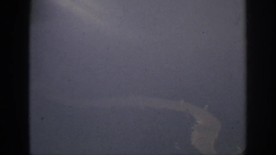 1962 Anaheim-area 8mm frame showing a hazy aircraft-window view of pale water or shoreline against darker terrain.