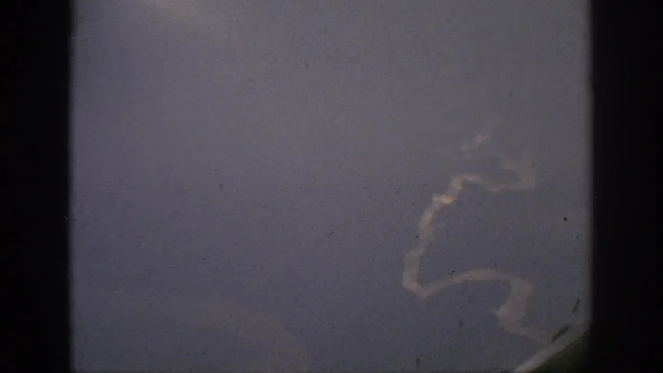 1962 Anaheim-area 8mm aerial view with a curved pale water or shoreline form visible through dark aircraft-window framing.
