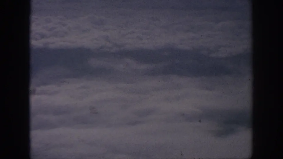 Archival 8mm view from an airplane window above dense clouds, with muted color and visible film texture.