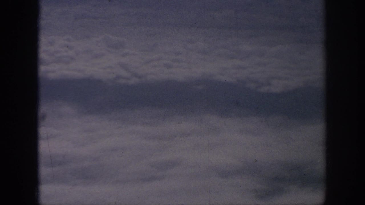 1962 8mm home-movie view from an airplane window, showing layered clouds beyond dark cabin framing.