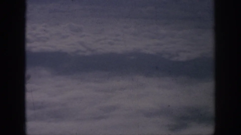 1962 8mm home-movie view from an airplane window, showing layered clouds beyond dark cabin framing.
