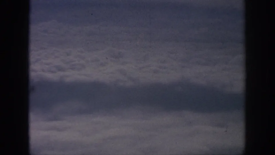 Airplane-window view from the 1962 travel footage, with clouds filling the frame behind dark vertical cabin framing.