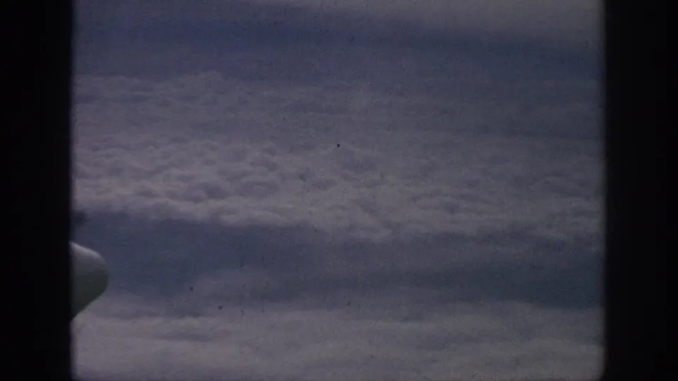 Vintage 8mm airplane-window view with clouds below and a small wing or engine shape visible near the lower left.