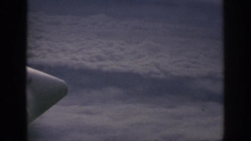 1962 8mm travel frame showing an airplane wing or engine shape against layered clouds outside the window.