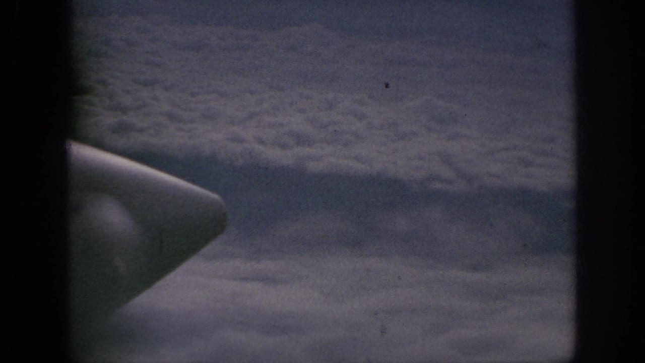1962 8mm home-movie view from an airplane window above clouds, with dark framing and pale aircraft structure at left.