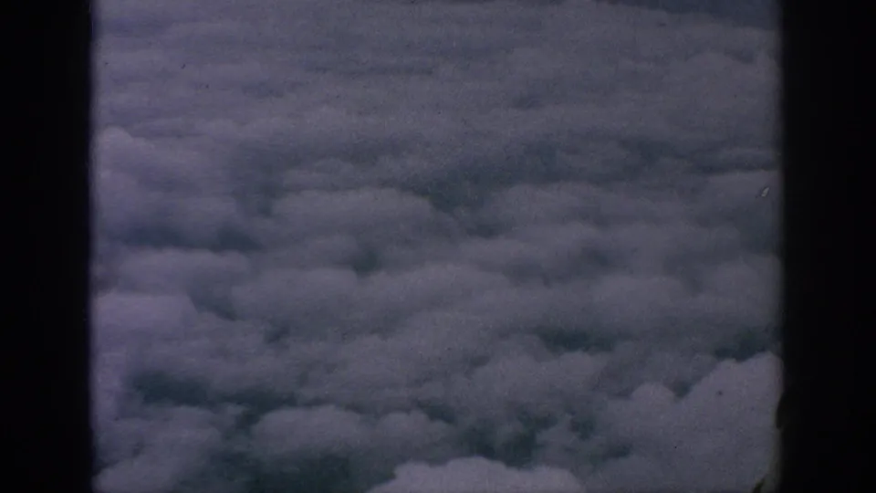 1962 8mm passenger-window view looking down at dense cloud cover, bordered by the dark aircraft window frame.