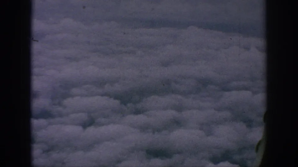 Aerial cloud view from the 1962 home-movie clip, framed by dark aircraft-window or film-gate edges.