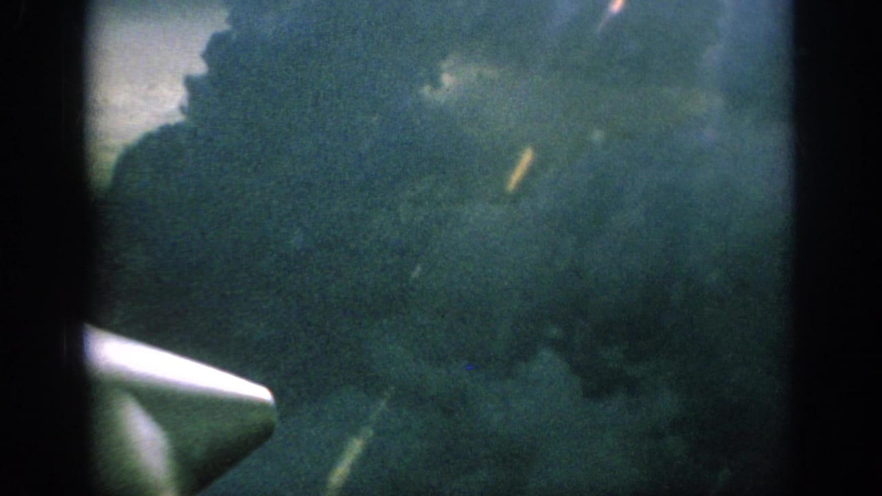 1962 home-movie frame looking past a white aircraft propeller or wingtip toward dense blue-green cloud cover.