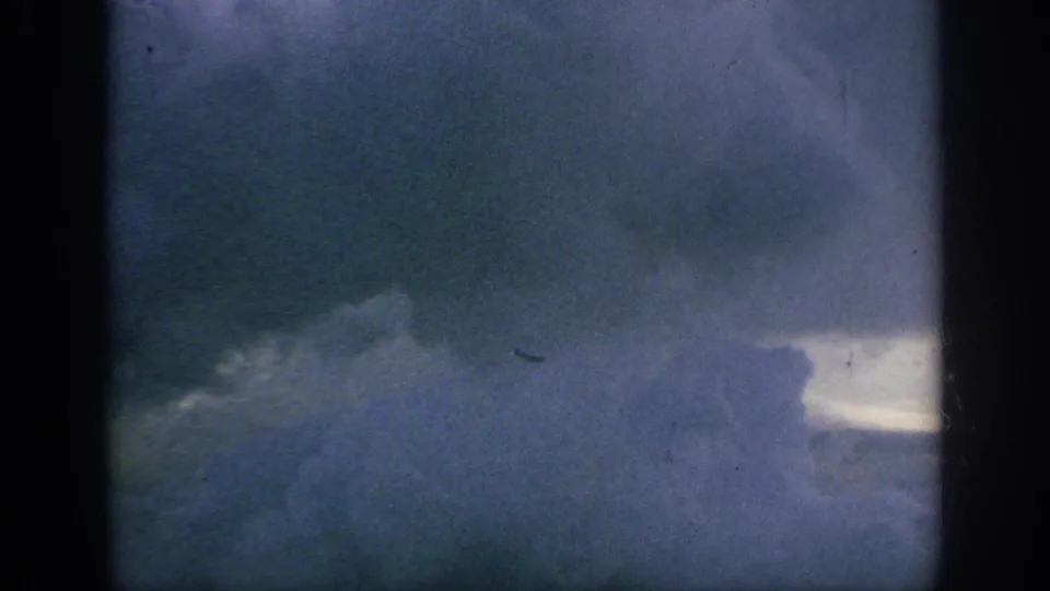 1962 home-movie airplane-window frame showing blue-gray clouds, a pale cloud break, and dark film-frame borders.