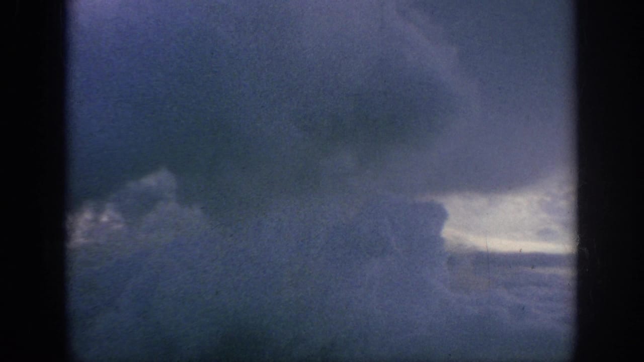 1962 8mm home-movie view of heavy blue-gray clouds through dark aircraft-window edges, with soft grain and fading.