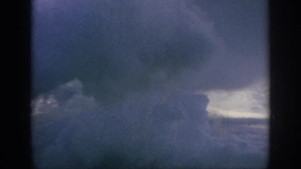 1962 8mm home-movie view of heavy blue-gray clouds through dark aircraft-window edges, with soft grain and fading.