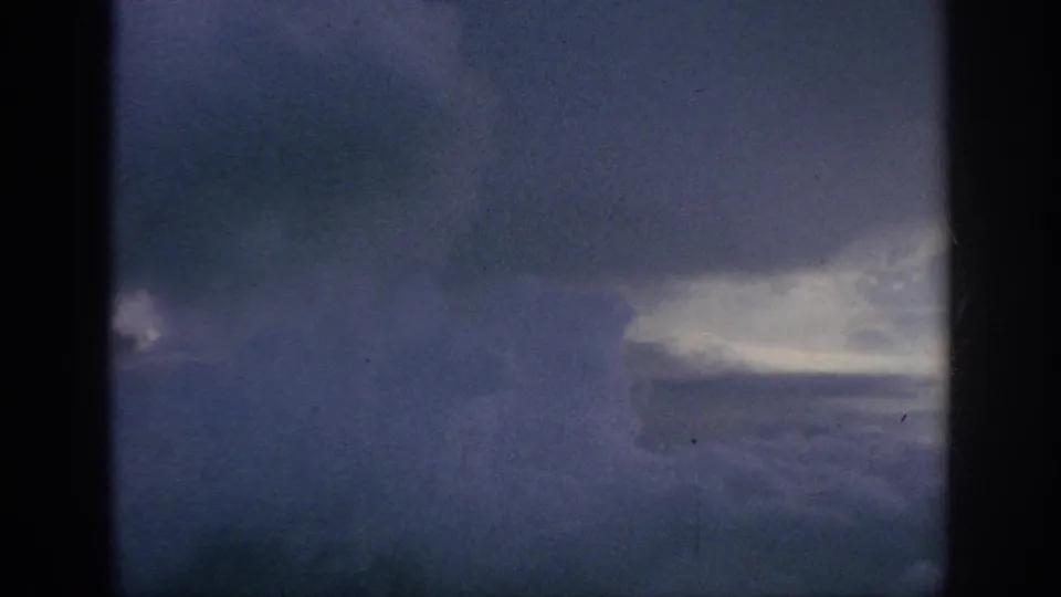 1962 aerial home-movie view of blue-gray cloud cover with a bright opening, framed by dark window edges.