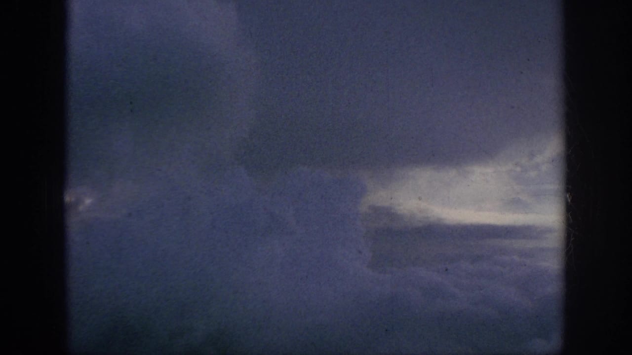 1962 8mm home-movie view of blue-gray clouds and sky seen from an airplane window, with dark frame edges and film grain.