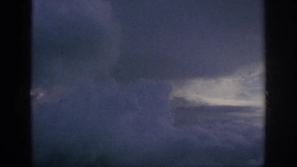 1962 8mm home-movie view of blue-gray clouds and sky seen from an airplane window, with dark frame edges and film grain.