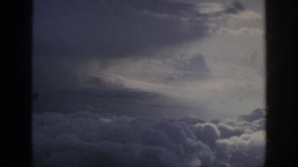 1962 8mm airplane-window view of layered clouds, hazy light, and dark frame borders with visible film texture.
