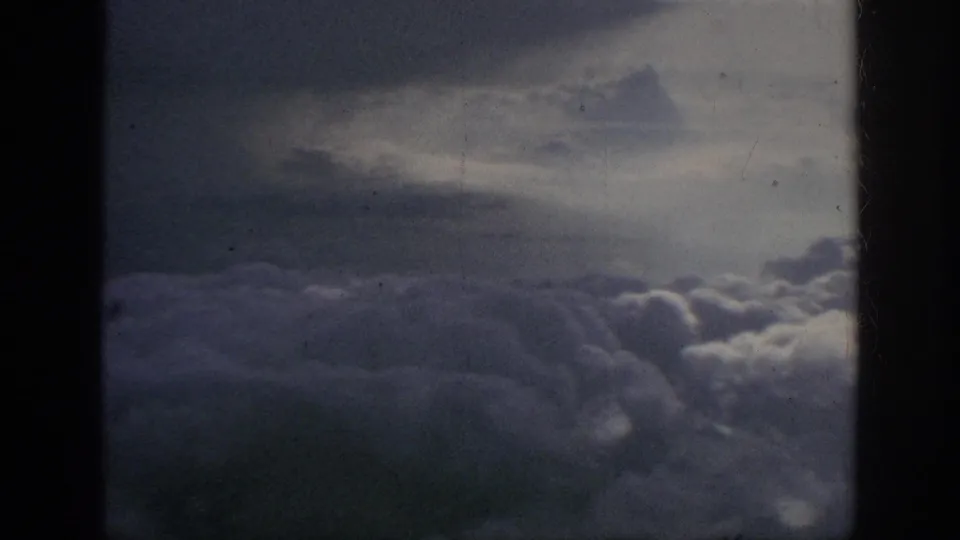 1962 8mm home-movie aerial view of dense cloud banks below a muted sky, bordered by dark window or gate edges.