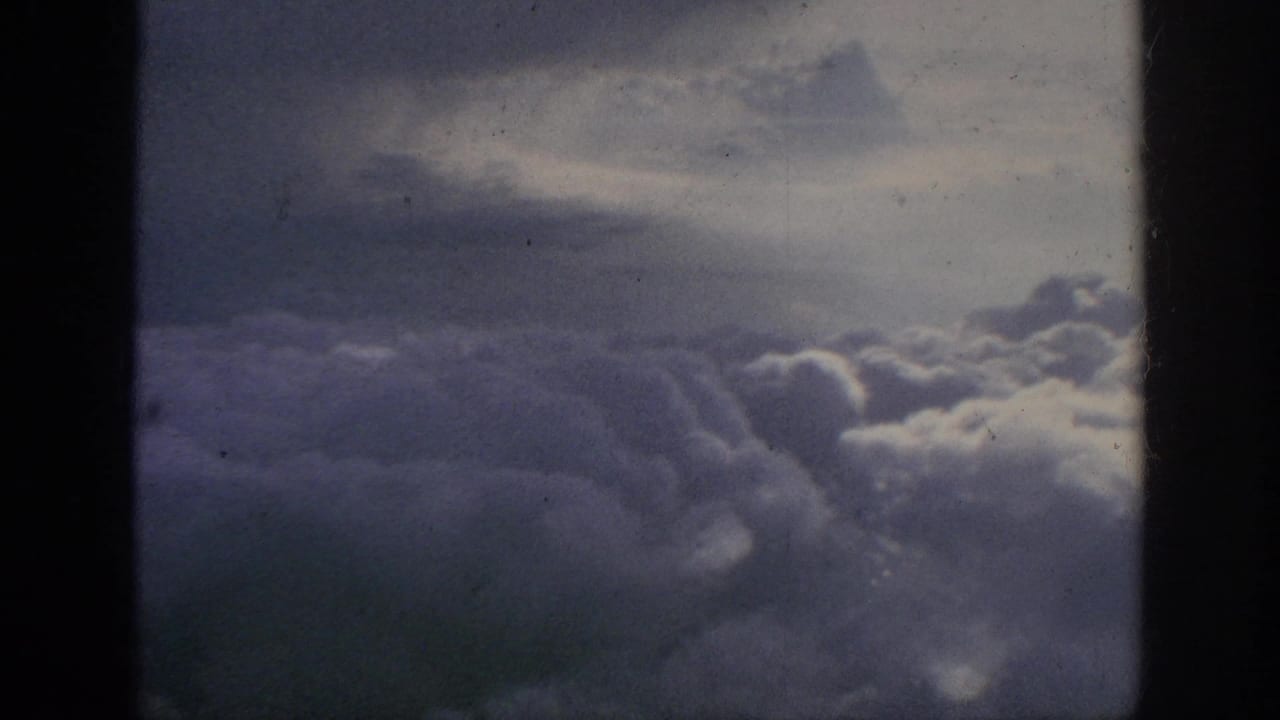 1962 8mm passenger-window view of thick clouds below a muted sky, bordered by dark aircraft-window edges.