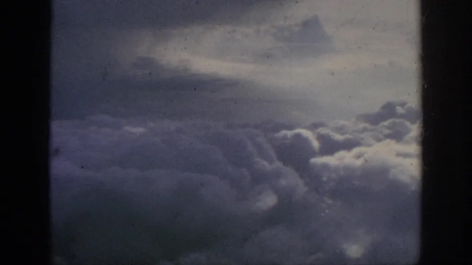 1962 8mm passenger-window view of thick clouds below a muted sky, bordered by dark aircraft-window edges.