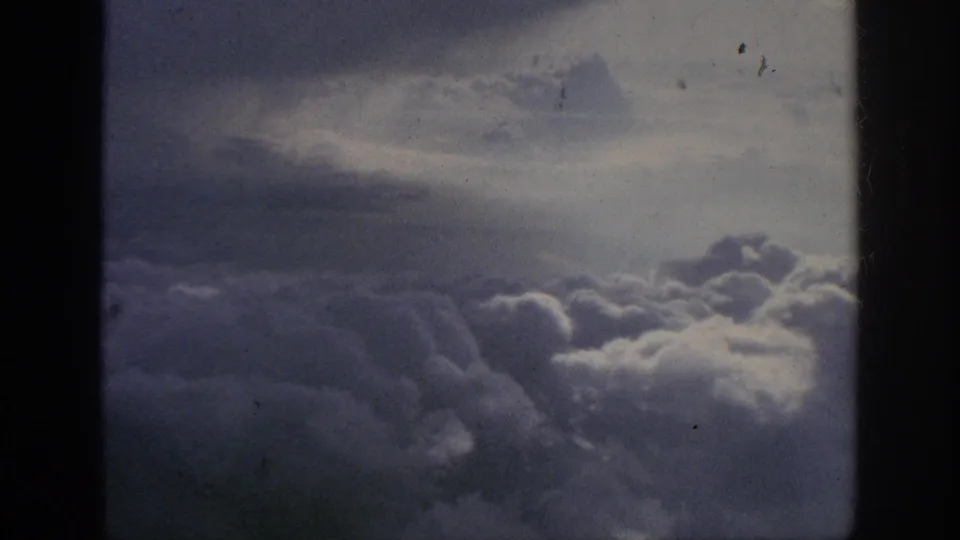 Vintage 8mm view from inside an airplane, showing fluffy cloud layers and a subdued sky through the window.