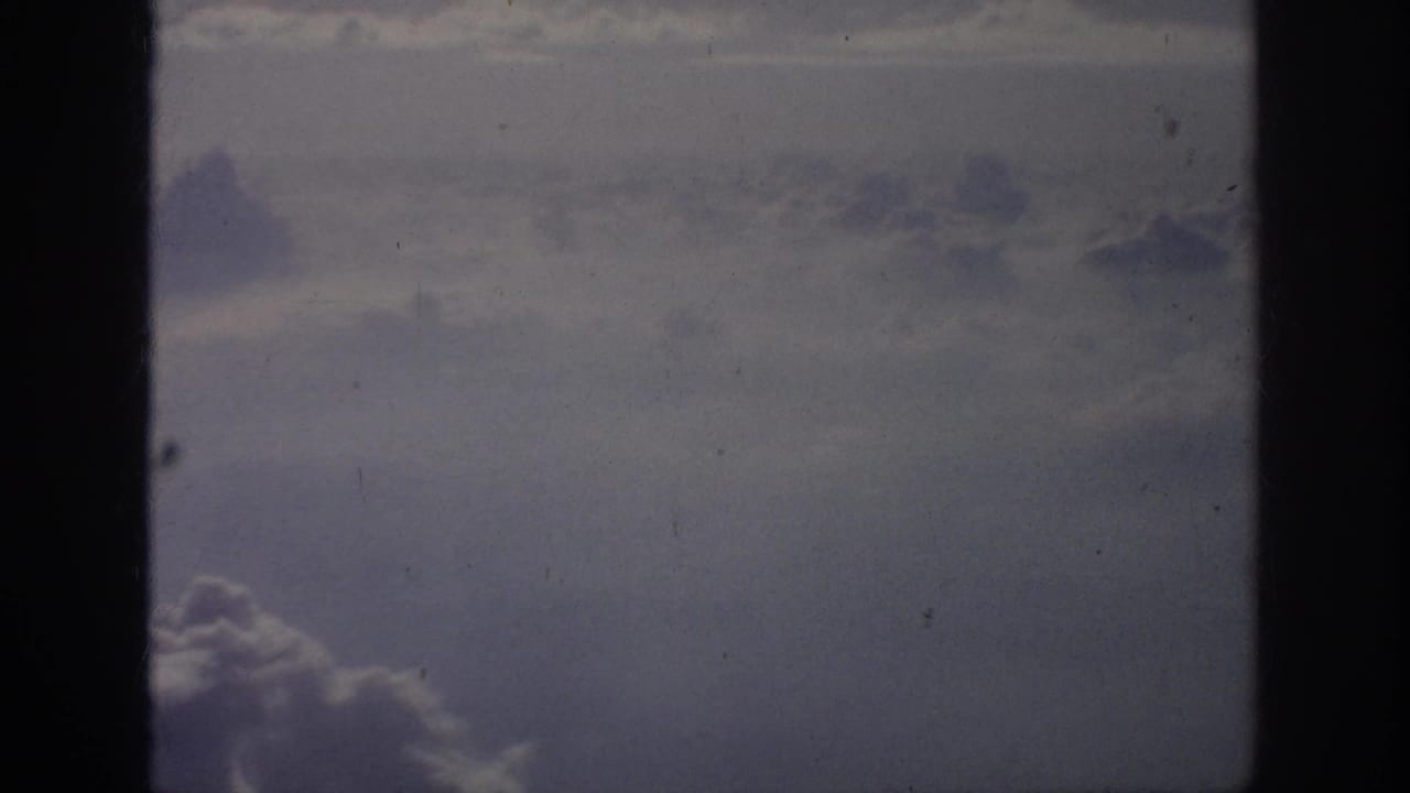 1962 8mm home-movie view of cloud layers through a dark aircraft window frame, with soft blue-gray color and film grain.