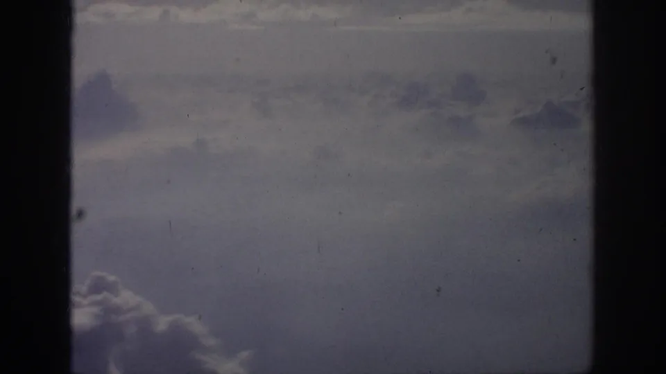1962 8mm home-movie view of cloud layers through a dark aircraft window frame, with soft blue-gray color and film grain.
