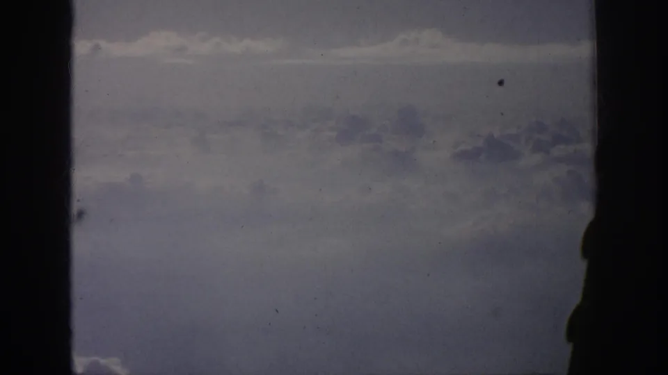 Aged 8mm frame showing a subdued sky and cloud bank through an aircraft window, with a central dark frame bar.