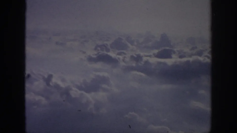 1962 home-movie aerial view of thicker cloud cover below the window line, tinted blue-purple by aged 8mm film.