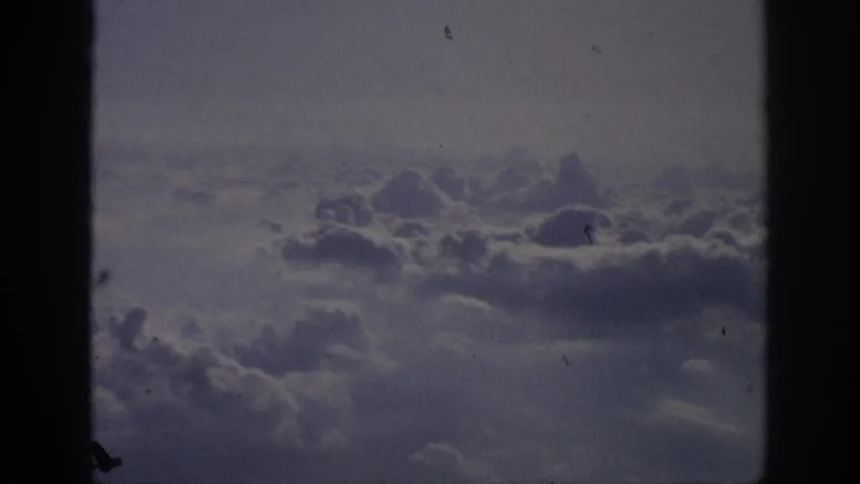 Soft 8mm aircraft-window view of layered clouds and muted sky, with dark framing and visible film wear.