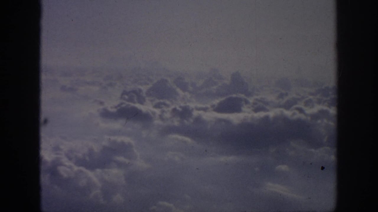 1962 8mm home-movie view of layered clouds through a left-side airplane window, with soft grain and muted color.
