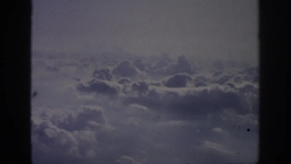1962 8mm home-movie view of layered clouds through a left-side airplane window, with soft grain and muted color.