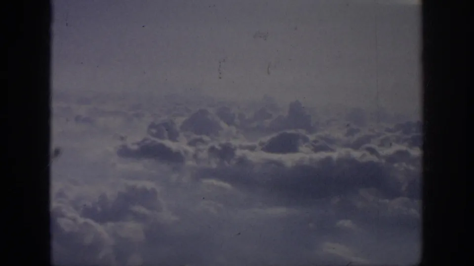 Aerial cloudscape from the supplied 1962 travel footage, seen through dark airplane window framing.