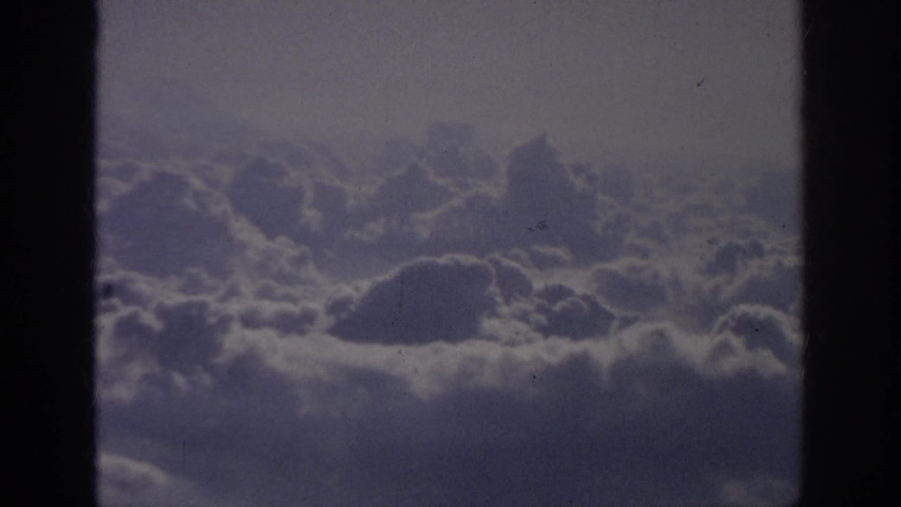 1962 8mm home-movie frame showing soft cloud banks viewed from an airplane-window perspective, with dark borders and visible film grain.