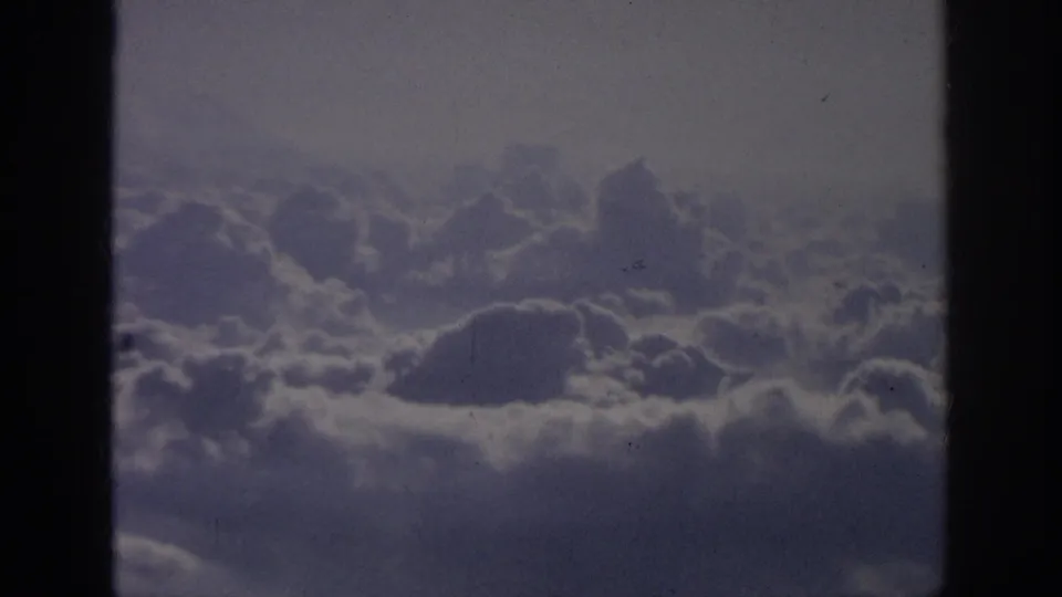 1962 8mm home-movie frame showing soft cloud banks viewed from an airplane-window perspective, with dark borders and visible film grain.