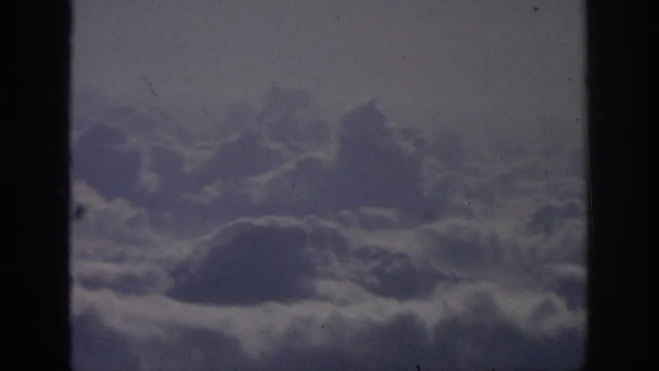A muted 1962 8mm frame of layered clouds from an aerial viewpoint, with faded color, grain, and dark framing at the sides.